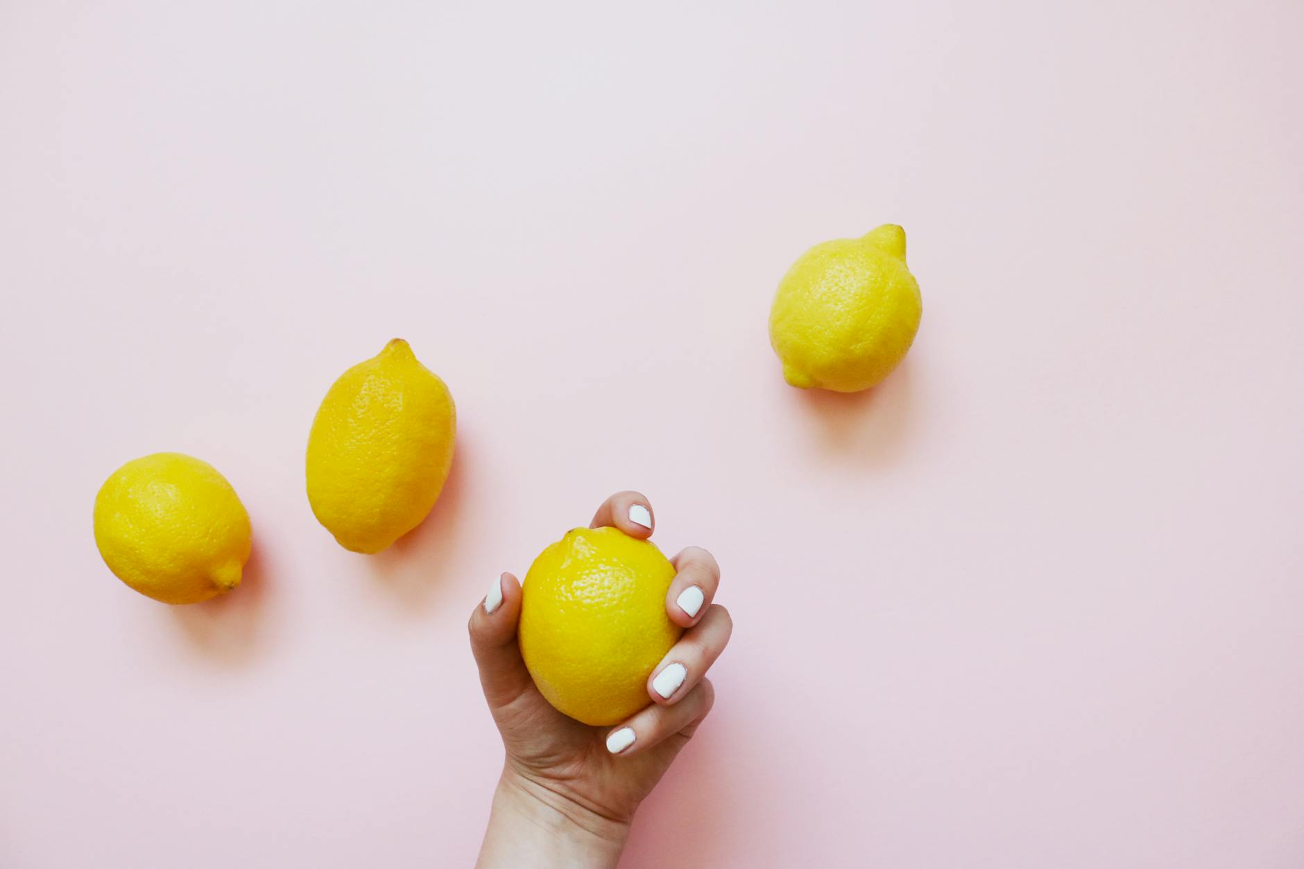 Una mano con unghie bianche che tiene un limone su uno sfondo rosa morbido, circondata da tre limoni aggiuntivi.