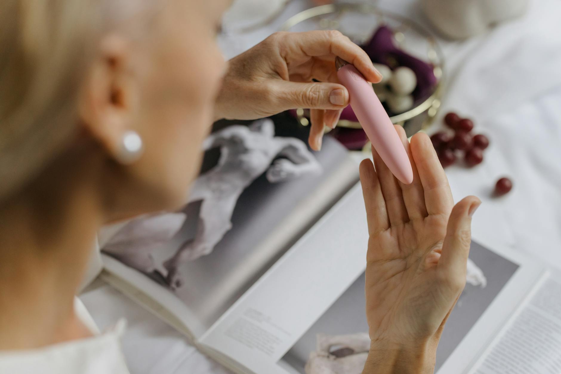 Close-up of a woman holding a pink vibrator, with art books in the background, emphasizing intimate and personal themes.